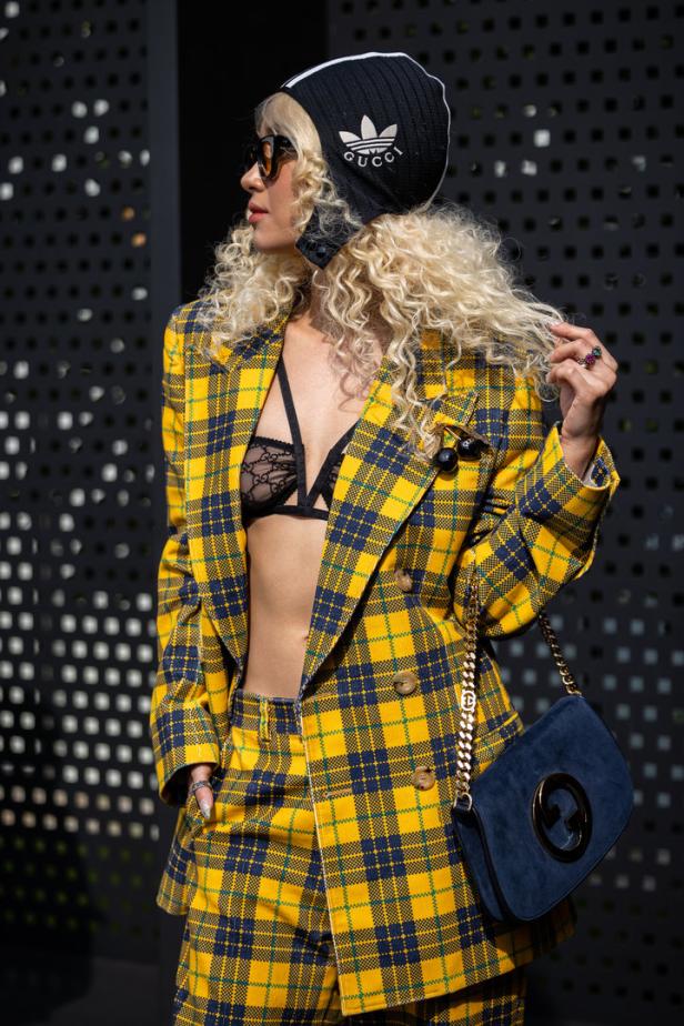 Eine Frau mit blonden Locken trägt einen gelb-blau karierten Anzug, eine Mütze und eine blaue Handtasche.