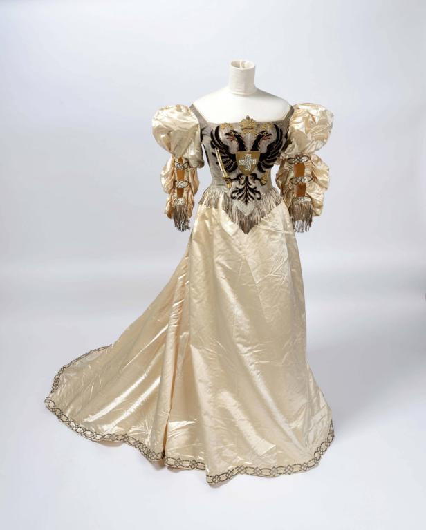Ein cremefarbenes Ballkleid mit Puffärmeln und einem aufwendigen, heraldischen Wappen auf dem Oberteil.