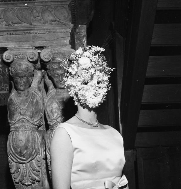 Eine Frau mit einem Blumenhut, der ihr Gesicht verdeckt, steht vor Statuen.