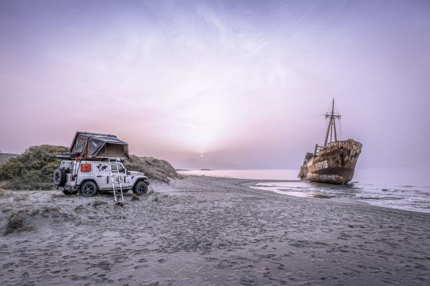 Ein Jeep mit Dachzelt steht am Strand, im Hintergrund ein altes Schiffswrack.