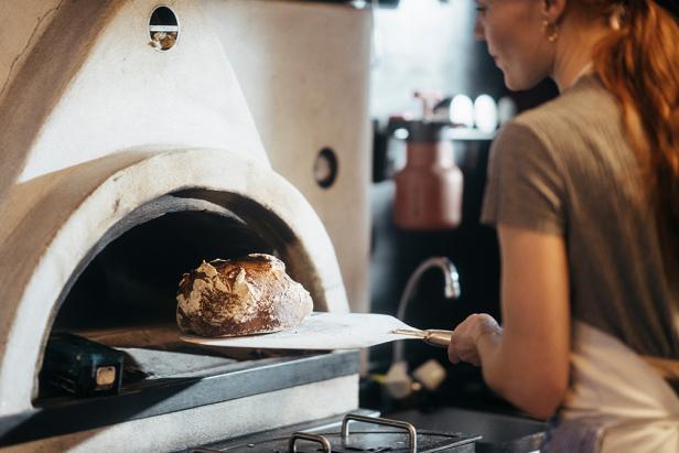 Eine Frau schiebt ein Brot mit einem Brotschieber in einen Steinofen.