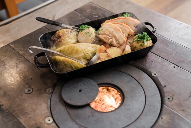 Ein Bräter mit Schweinebraten, Knödeln und Kraut steht auf einem Holzofen.