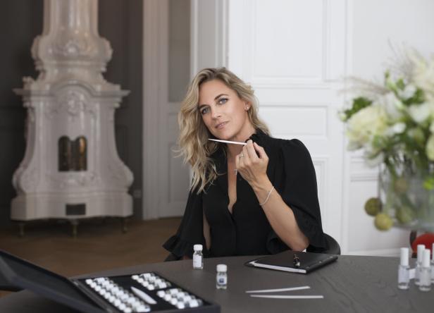 Eine blonde Frau in einem schwarzen Kleid testet einen Duftstreifen an einem Tisch mit Parfümflaschen.