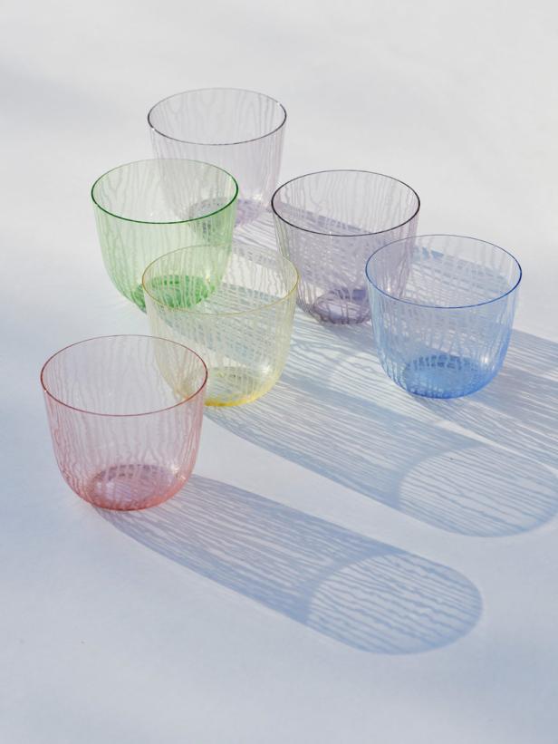 Sechs bunte Trinkgläser stehen auf einer hellen Oberfläche und werfen Schatten.