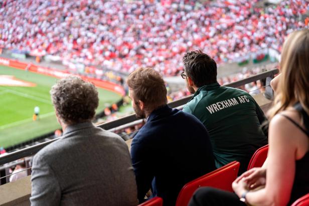 Zuschauer, darunter einer mit einer „Wrexham AFC“-Jacke, beobachten ein Fußballspiel.