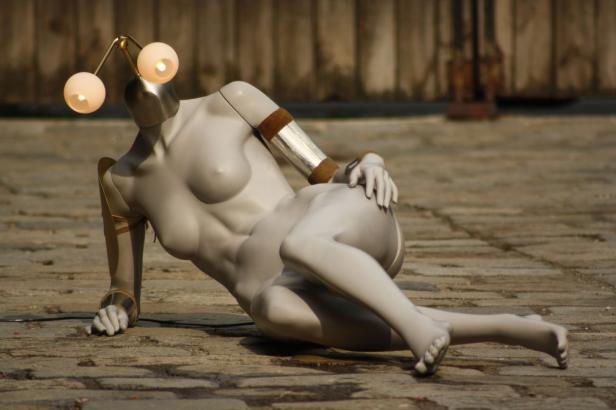 Eine stilisierte weibliche Figur mit Lampen als Kopf liegt auf einem Steinboden.