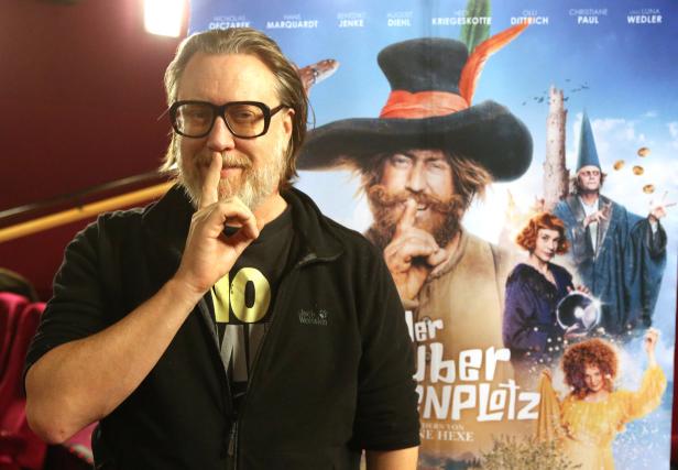 Ein Mann mit Brille hält den Finger vor den Mund, vor einem Filmplakat für „Der Teufel vom Mühlenplatz“.