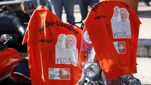 Zwei orangefarbene T-Shirts mit Bildern von Papst Franziskus hängen an einem Harley-Davidson-Motorrad.