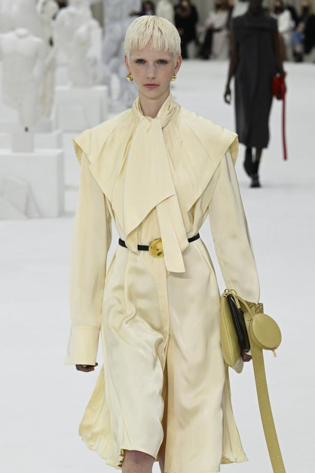 Ein Model präsentiert ein helles Kleid mit passender Handtasche auf dem Laufsteg.