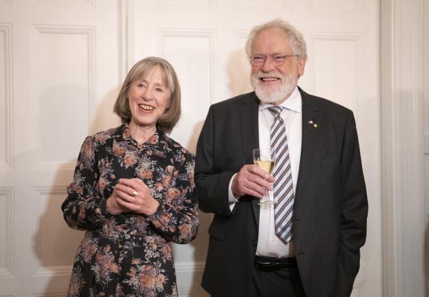 Ein älteres Paar steht lächelnd vor einer weißen Tür, der Mann hält ein Glas Sekt.