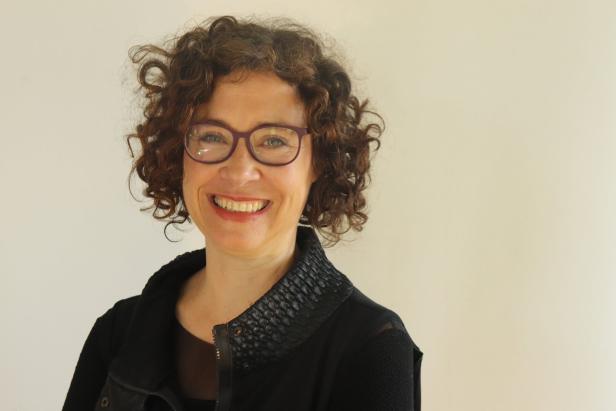 Eine lächelnde Frau mit lockigem Haar und Brille vor einem hellen Hintergrund.