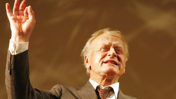 Ein älterer Mann im Anzug hebt die Hand.