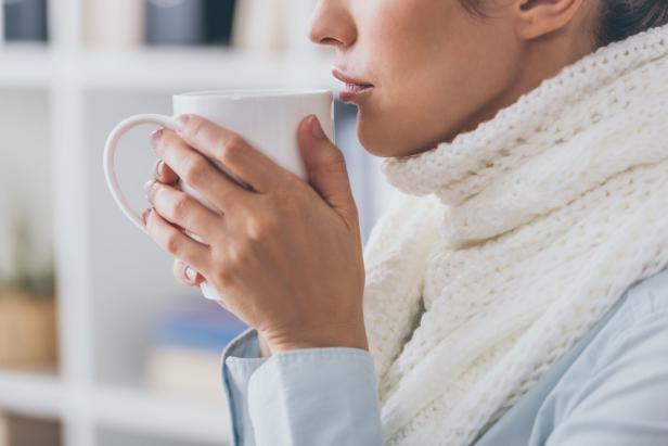 Eine Frau mit einem weißen Schal trinkt aus einer weißen Tasse.