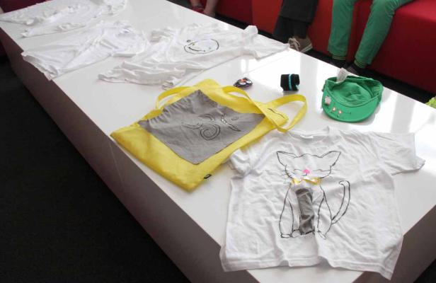 Auf einem Tisch liegen mehrere T-Shirts und eine gelbe Stofftasche mit verschiedenen Motiven.