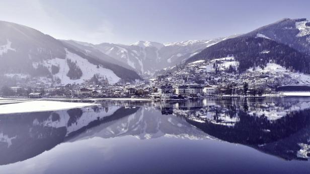 Ein verschneites Dorf liegt am Ufer eines Sees, umgeben von Bergen.