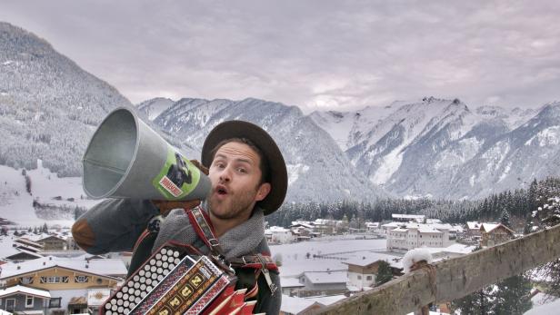Ein Mann mit Hut und Akkordeon ruft durch ein Megaphon vor einer verschneiten Berglandschaft.