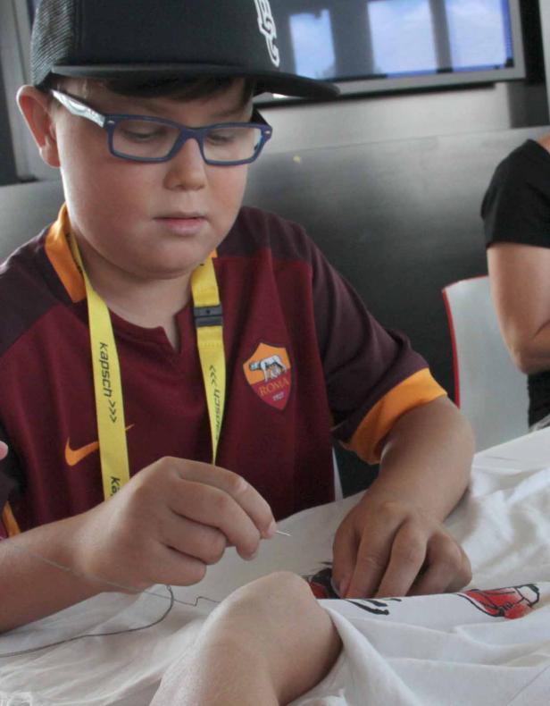 Ein Junge mit Brille und AS Rom Trikot näht etwas auf einem weißen Stoff.
