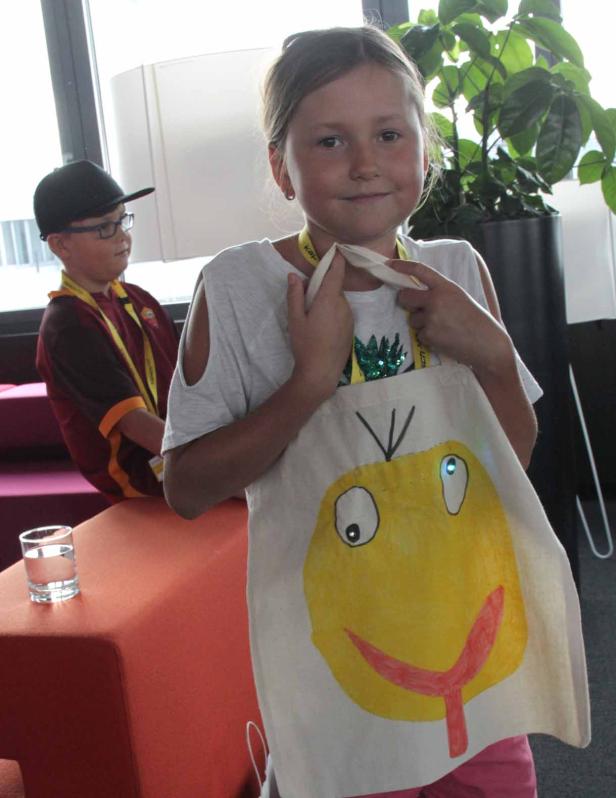 Ein Mädchen präsentiert eine Stofftasche mit einem gelben, gemalten Gesicht darauf.