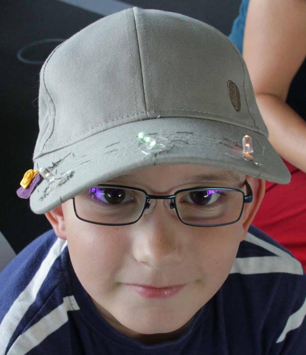 Ein Junge mit Brille und einer grauen Kappe, die mit Lichtern und einer Blume verziert ist.
