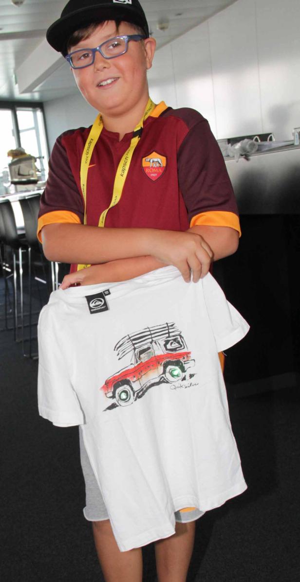 Ein Junge mit Brille hält ein T-Shirt mit einem Autoaufdruck in der Hand.