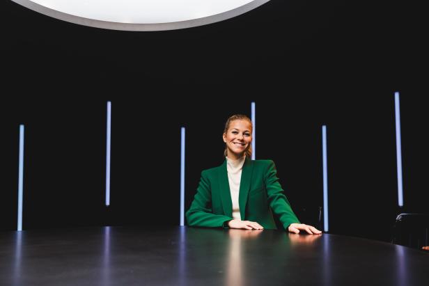 Eine Frau in einem grünen Blazer sitzt an einem runden Tisch vor einem schwarzen Hintergrund.