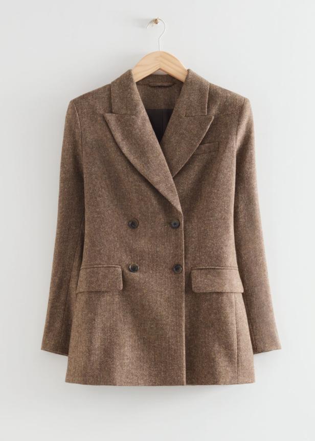 Ein brauner Blazer mit doppelter Knopfleiste hängt an einem Holzbügel vor einer weißen Wand.