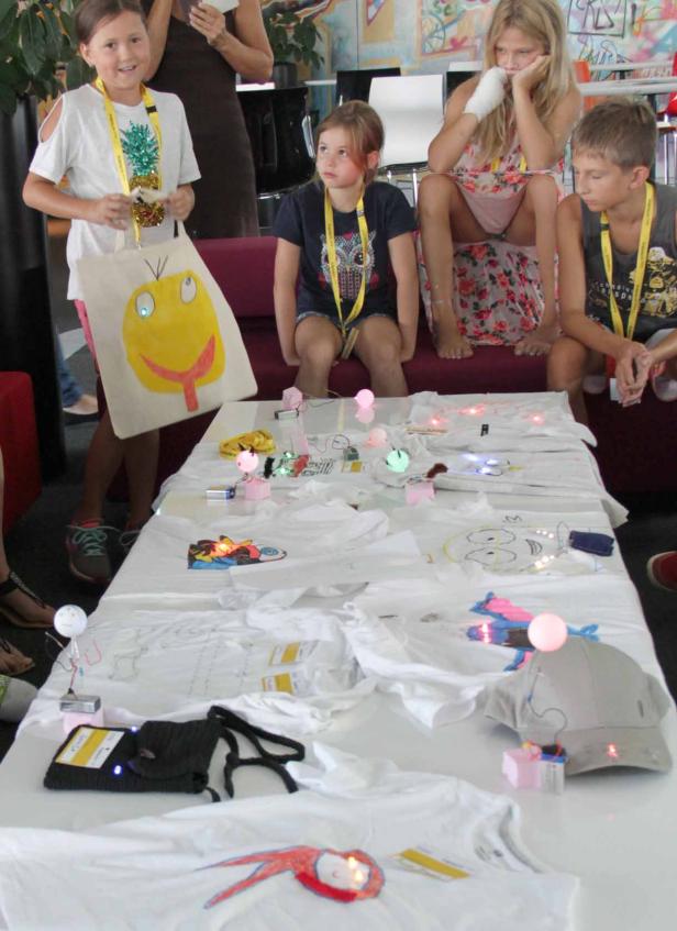 Kinder sitzen an einem Tisch mit selbstgestalteten, leuchtenden T-Shirts und Taschen.