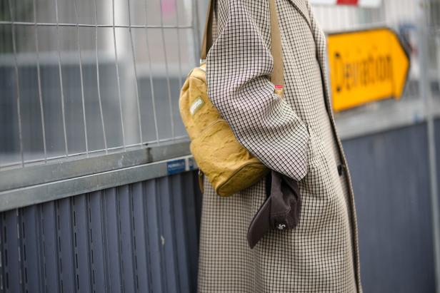 Eine Person mit einem karierten Mantel und einem gelben „Supreme“-Rucksack steht an einem Bauzaun.