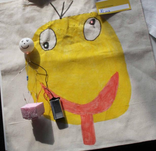 Eine Stofftasche mit einer gelben, kindlichen Monsterzeichnung und einer kleinen elektronischen Schaltung.