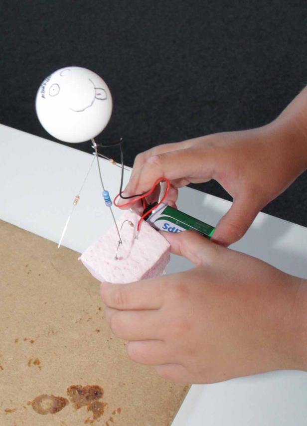 Ein Kind hantiert mit einer Batterie und einem Schaltkreis, an dem ein Styroporkopf befestigt ist.