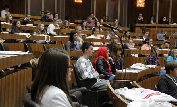 Zahlreiche Jugendliche sitzen in einem großen Saal mit Holzbänken und Tischen.