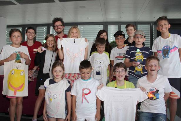 Eine Gruppe von Kindern und Erwachsenen präsentiert stolz ihre selbstgestalteten T-Shirts mit leuchtenden Elementen.