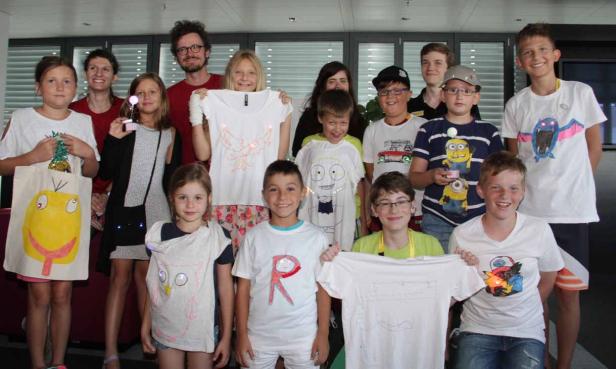 Eine Gruppe Kinder und Erwachsene präsentiert stolz ihre selbstgestalteten T-Shirts und Taschen mit leuchtenden Elementen.