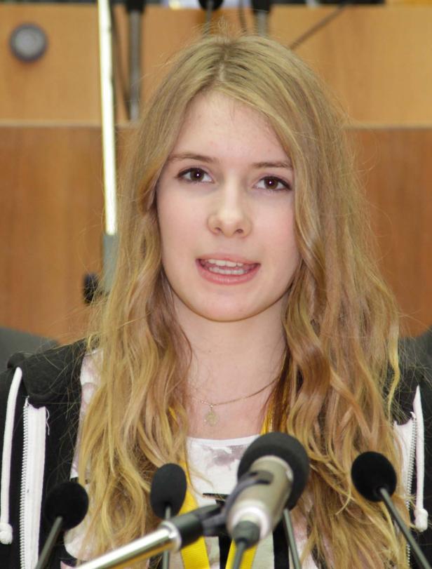 Eine junge Frau mit blonden, gewellten Haaren spricht in mehrere Mikrofone.