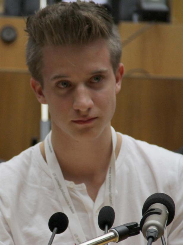 Ein junger Mann mit blonden Haaren steht vor mehreren Mikrofonen.