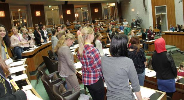 Eine Gruppe junger Leute steht in einem Sitzungssaal mit hölzernen Tischen und Stühlen.