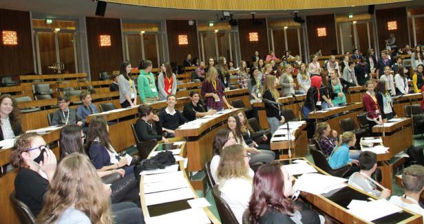 Eine Gruppe junger Leute sitzt in einem großen Saal mit Holzbänken und Tischen.