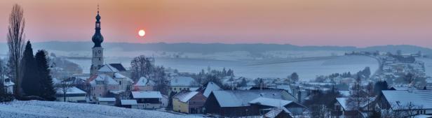 Winterliche Landschaft mit einer Kirche und schneebedeckten Häusern bei Sonnenaufgang.