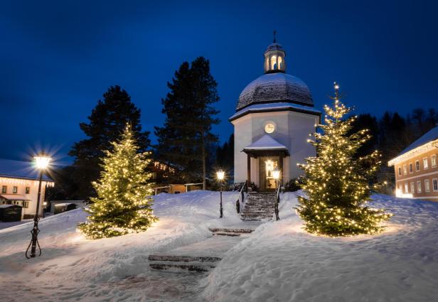 Eine kleine Kapelle im Schnee, geschmückt mit beleuchteten Weihnachtsbäumen.