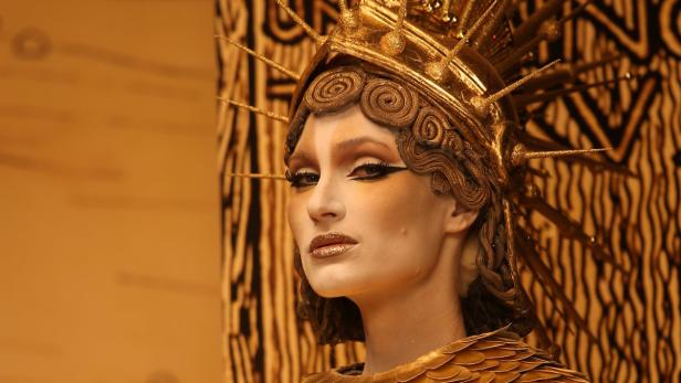 Eine Frau mit goldenem Kopfschmuck und Make-up vor einem gemusterten Hintergrund.