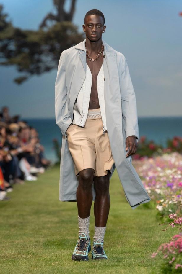 Ein dunkelhäutiger Mann in Shorts, Mantel und Sportschuhen läuft über einen Laufsteg.