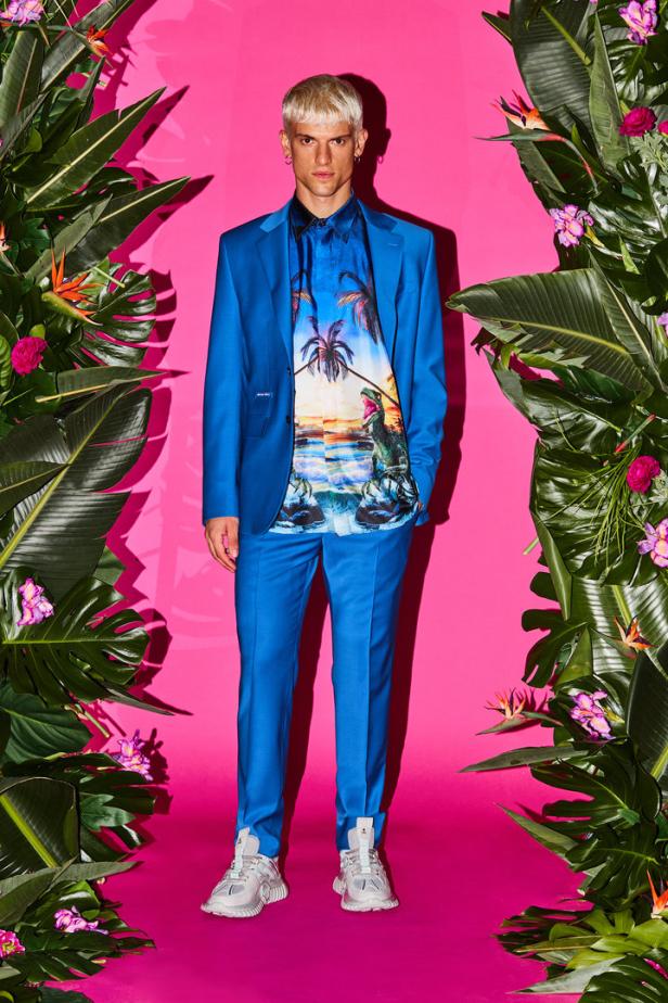 Ein Mann in einem blauen Anzug und einem Hemd mit Dinosaurier- und Palmenmuster vor einem rosa Hintergrund.