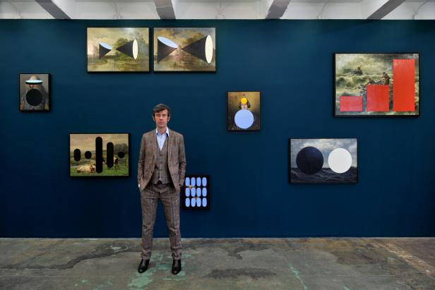 Ein Mann im Anzug steht vor einer blauen Wand mit mehreren Kunstwerken.