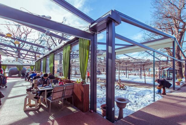 Ein Wintertag im Freien: Menschen sitzen in einem verglasten Café mit Blick auf einen verschneiten Park.