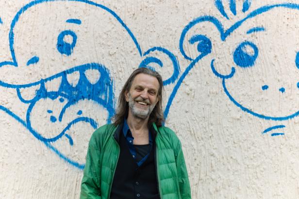 Ein lächelnder Mann steht vor einer Wand mit blauen Graffiti-Zeichnungen.