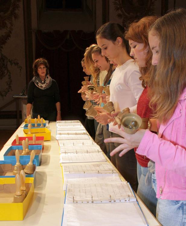 Eine Gruppe junger Frauen spielt mit Handglocken vor Notenblättern, unter der Leitung einer Dirigentin.