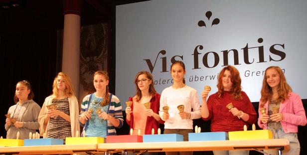 Eine Gruppe junger Frauen spielt auf der Bühne mit Handglocken vor dem „Vis fontis“-Logo.