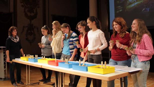 Eine Gruppe junger Frauen spielt Handglocken unter der Leitung einer Dirigentin.