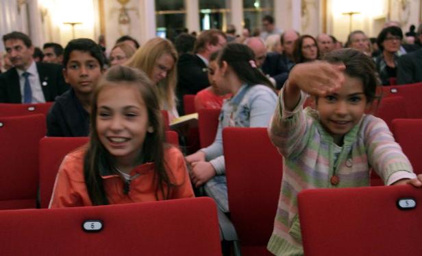 Kinder sitzen in einem Publikum auf roten Sitzen.