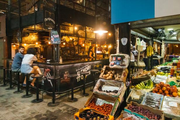 Ein Paar sitzt an der Bar eines Restaurants neben einem Marktstand mit Obst und Gemüse.